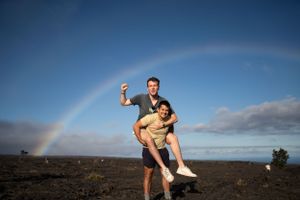 A couple poses playfully under a vibrant rainbow against a dramatic cloudy sky on volcanic terrain.