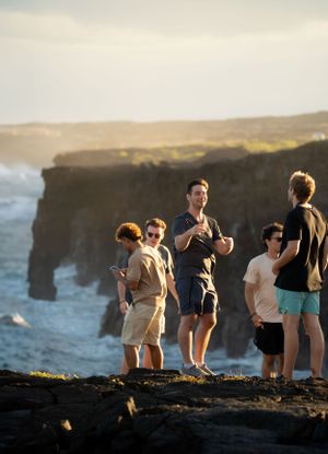 A group of tourists stand at the edge of dramatic ocean cliffs during sunset.