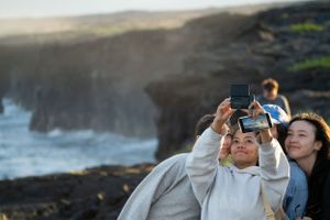 Tourists take photos at the edge of dramatic coastal cliffs with crashing waves below.