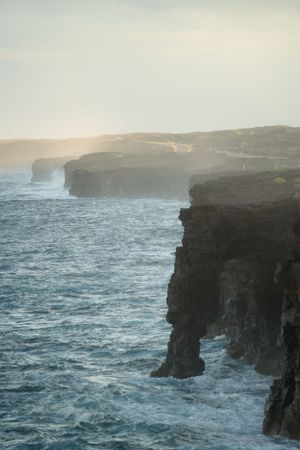 Dramatic volcanic cliffs rise above turbulent ocean waters during a misty sunset.