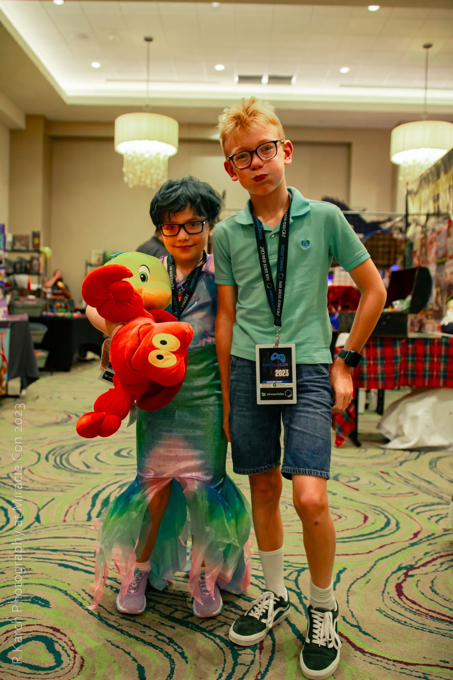 Two young attendees at a convention pose with an Elmo plush toy on a decorative patterned carpet.