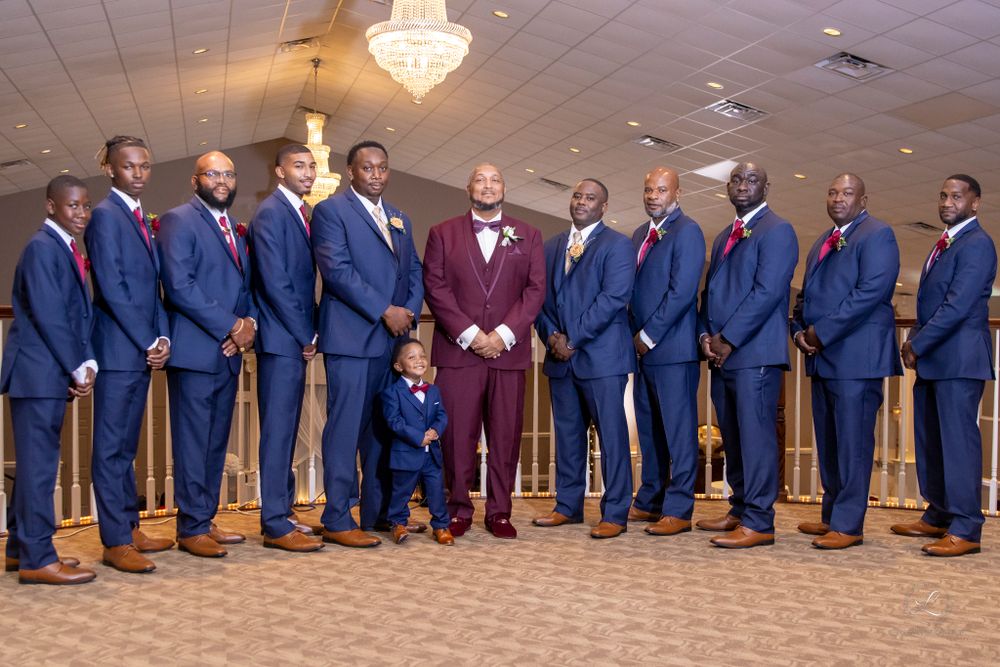 Wedding party in navy suits with groom in burgundy standing in formal reception venue.