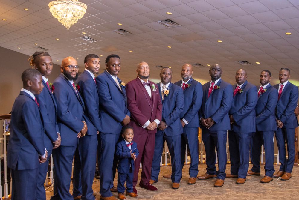 Large wedding party in navy suits and burgundy accents posing in front of crystal chandelier.