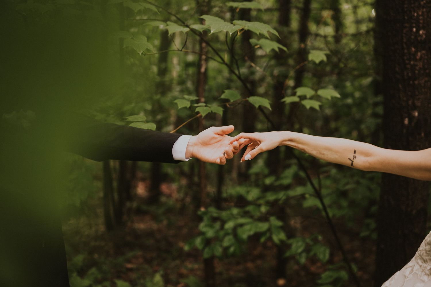 Bride and groom reach for each other's hands in a romantic moment in a lush green forest setting.