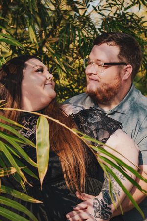 A couple embraces among lush green tropical plants, sharing a tender moment outdoors.