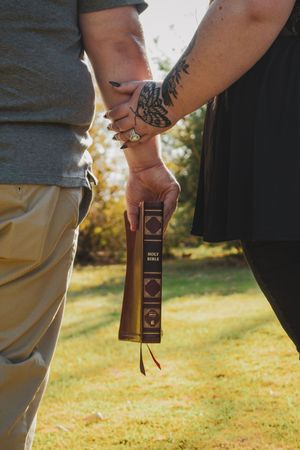 Couple holding hands and a Holy Bible outdoors, one with a tattoo and rings, standing on green grass in sunlight.