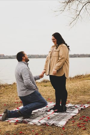 A couple sits on a picnic blanket by a lake during a romantic proposal moment with scattered red rose petals.