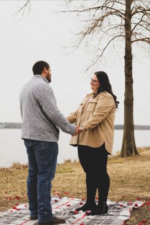 A couple shares a romantic proposal moment on a blanket by a lake with bare winter trees in the background.