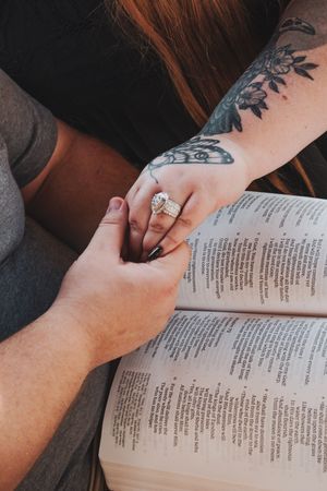 Couple holding hands over open Bible, woman wearing diamond ring with floral tattoo on forearm.