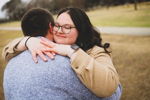 A couple in casual attire embraces lovingly in a grassy field during a golden hour photoshoot.