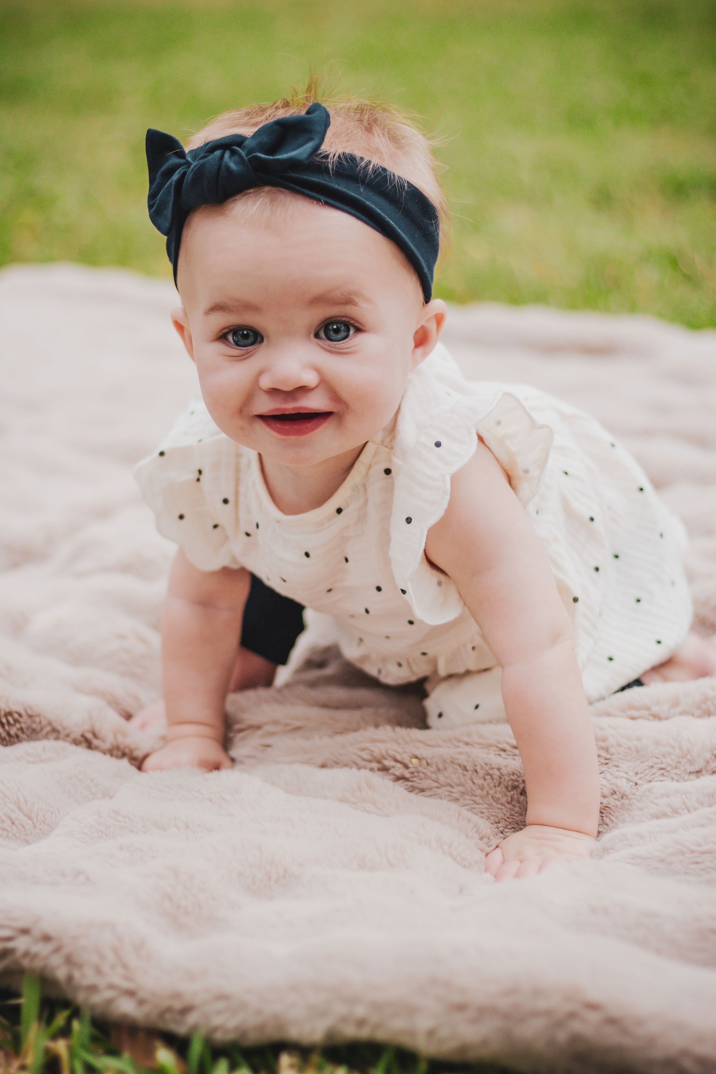 Smiling baby girl in white polka dot dress and black bow headband crawling on pink blanket outdoors.