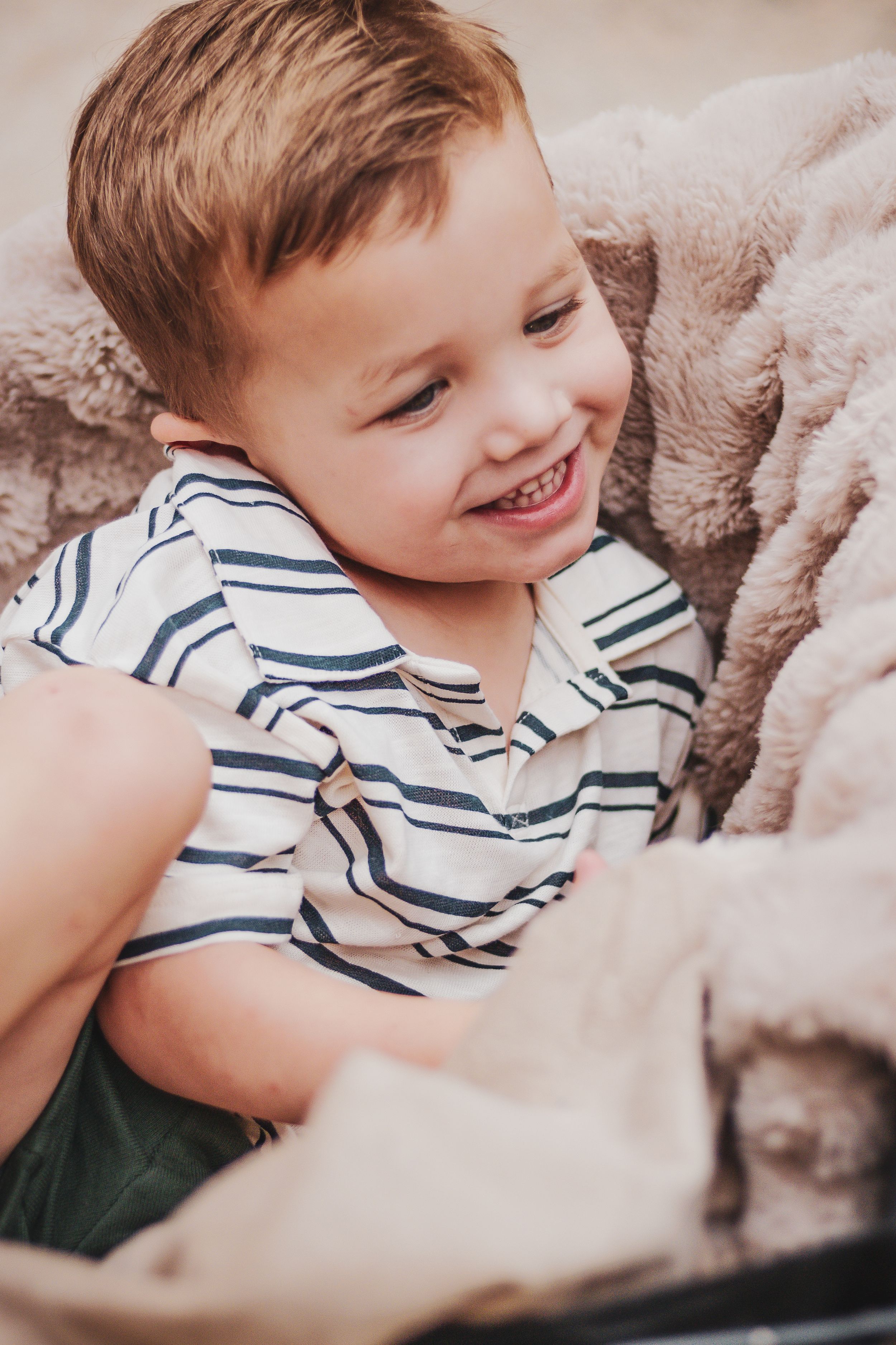 Smiling toddler boy in striped shirt relaxing on soft beige blanket, looking happy and playful.