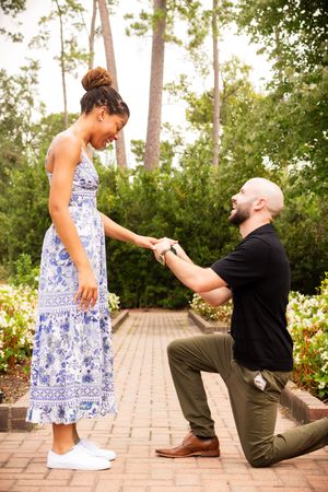 A romantic proposal moment on a brick path in a garden setting with tall trees and flowers in the background.