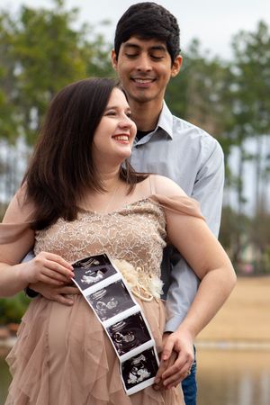 A couple in casual attire poses outdoors while holding ultrasound photos near a lake with pine trees in the background.
