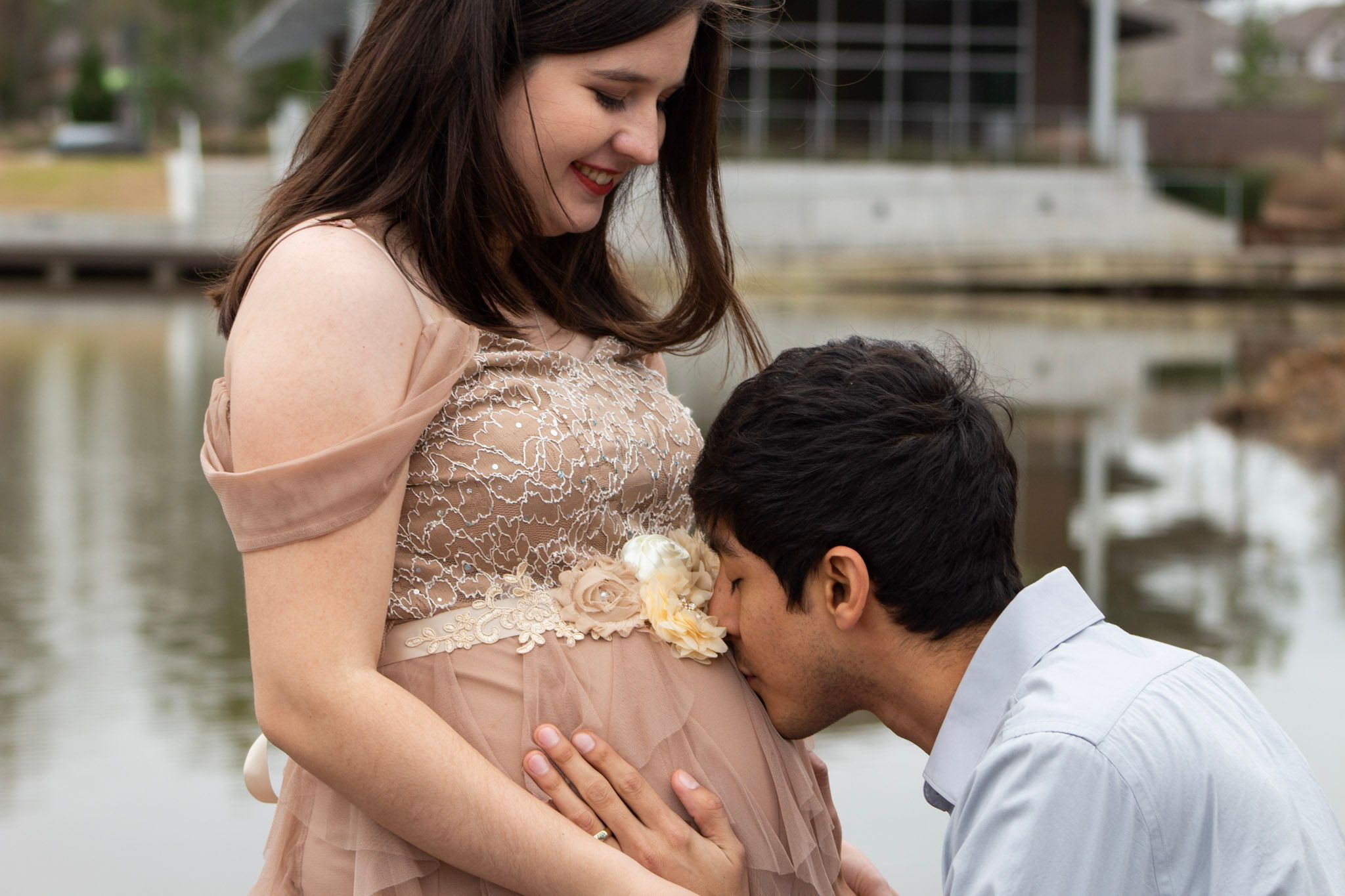 A couple shares an intimate maternity moment by a lake, with the partner kissing the pregnant belly in a tender pose.