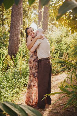 A couple embraces in a romantic moment among tall pine trees and sunlit foliage in a forest setting.