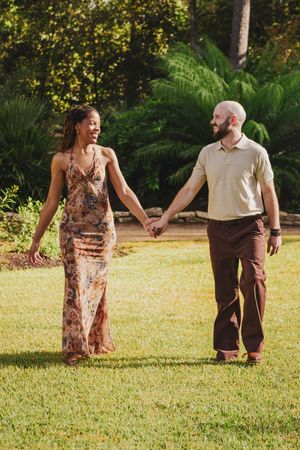 A couple holds hands while walking together on a sunny lawn with lush greenery in the background.