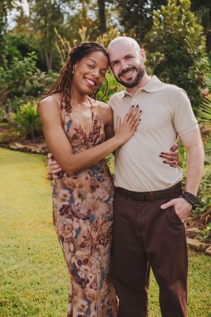 A couple embraces outdoors in a garden setting, with her wearing a floral dress and him in a light colored shirt.