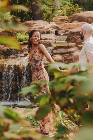 A couple shares a romantic moment near a cascading waterfall in a lush garden setting.