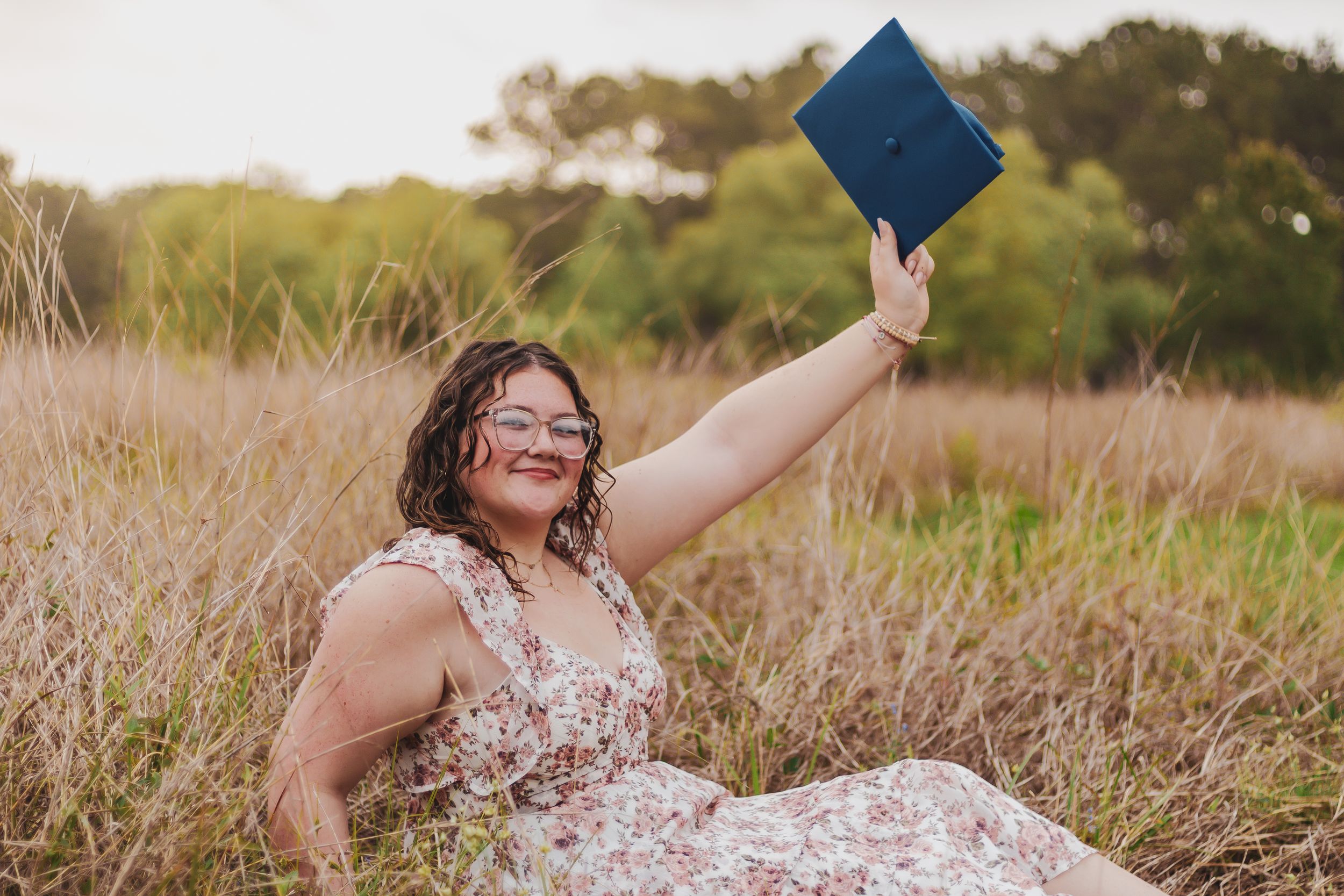 Woman in floral dress sitting in golden field, joyfully raising blue graduation cap in celebration.