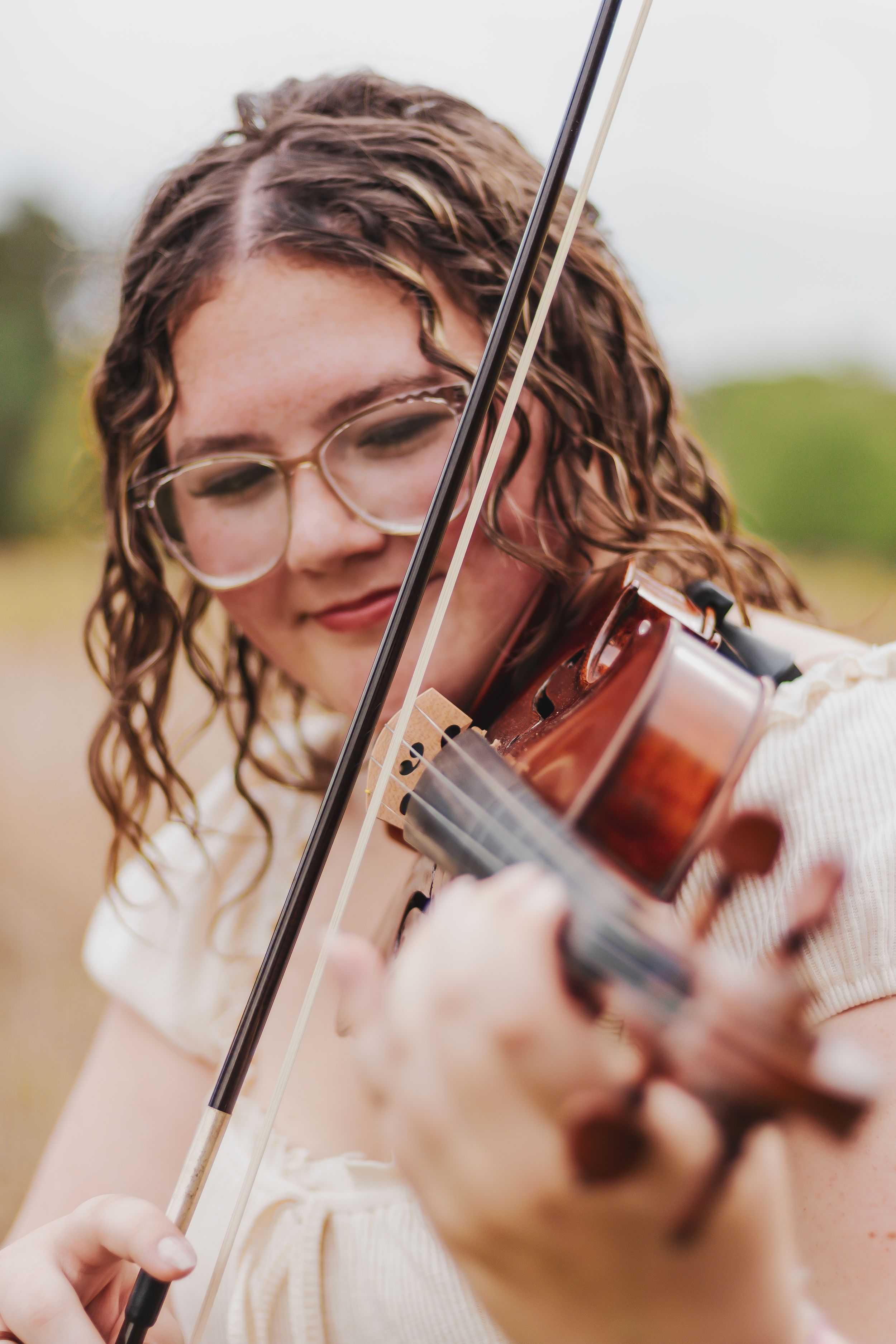 Young woman with curly hair and glasses playing violin outdoors in a field, focused on her music.