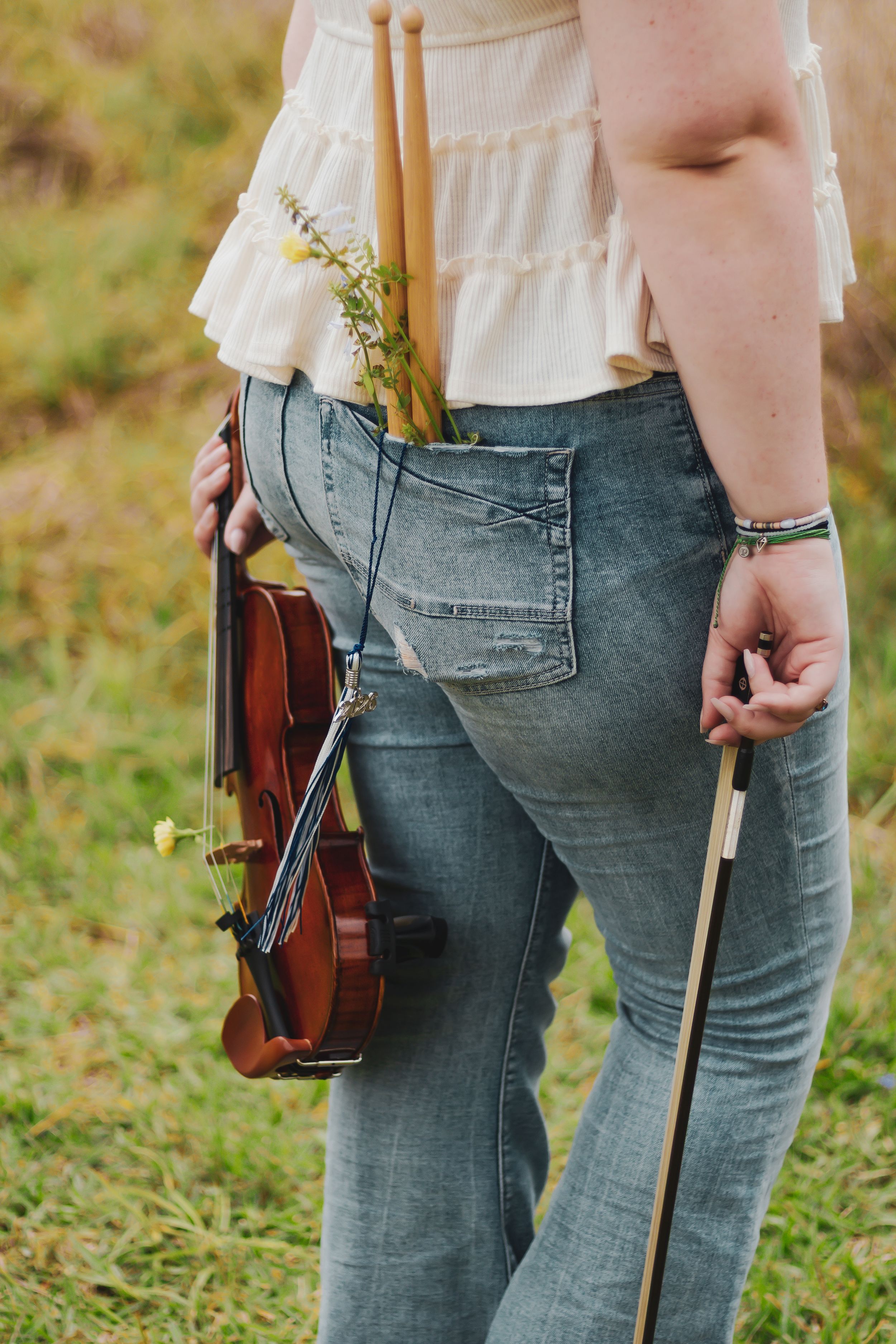 Woman in jeans holding a violin and bow with wildflowers tucked in her back pocket, standing in a field.