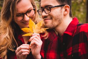 A couple wearing matching red plaid shirts share a laugh while holding yellow autumn leaves outdoors.