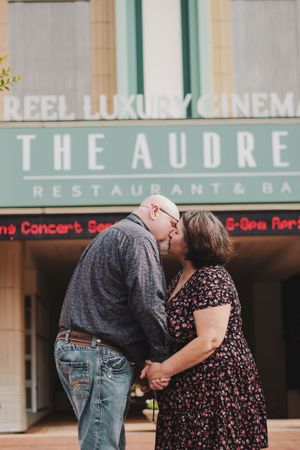 Couple sharing a romantic kiss in front of The Audrey Restaurant & Bar and Reel Luxury Cinema signs.