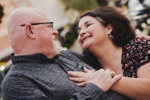 A smiling couple embraces outdoors, sharing a joyful moment, with the woman wearing a floral top and gold ring.