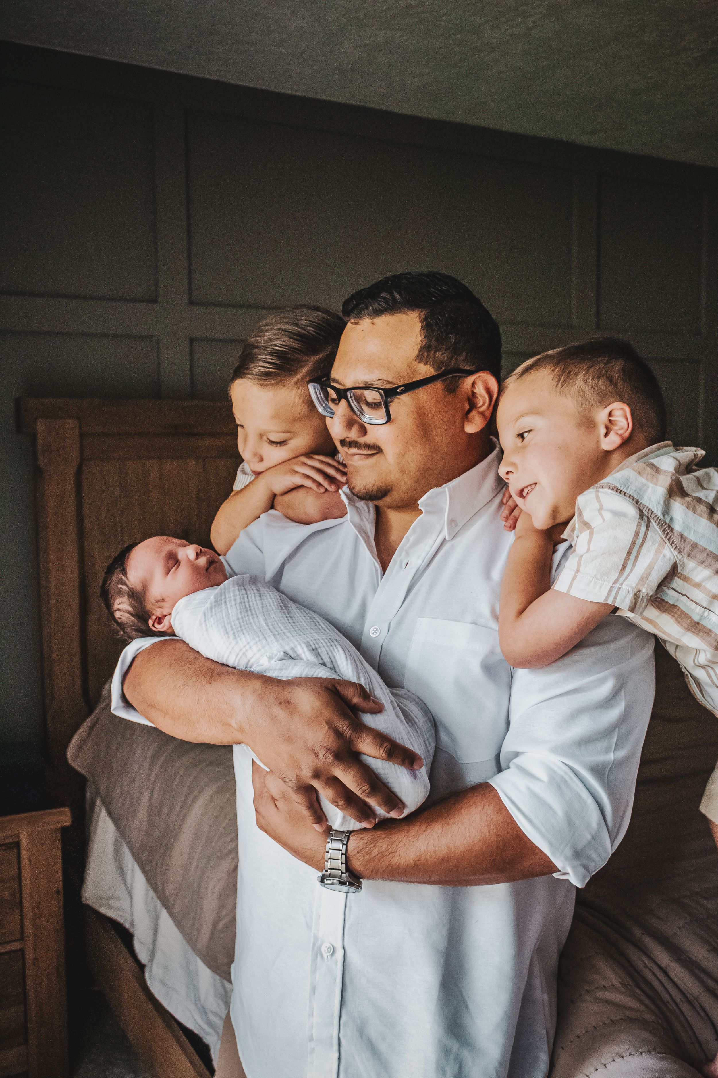 A father in glasses holds his newborn baby while two young boys lean on his shoulders indoors.