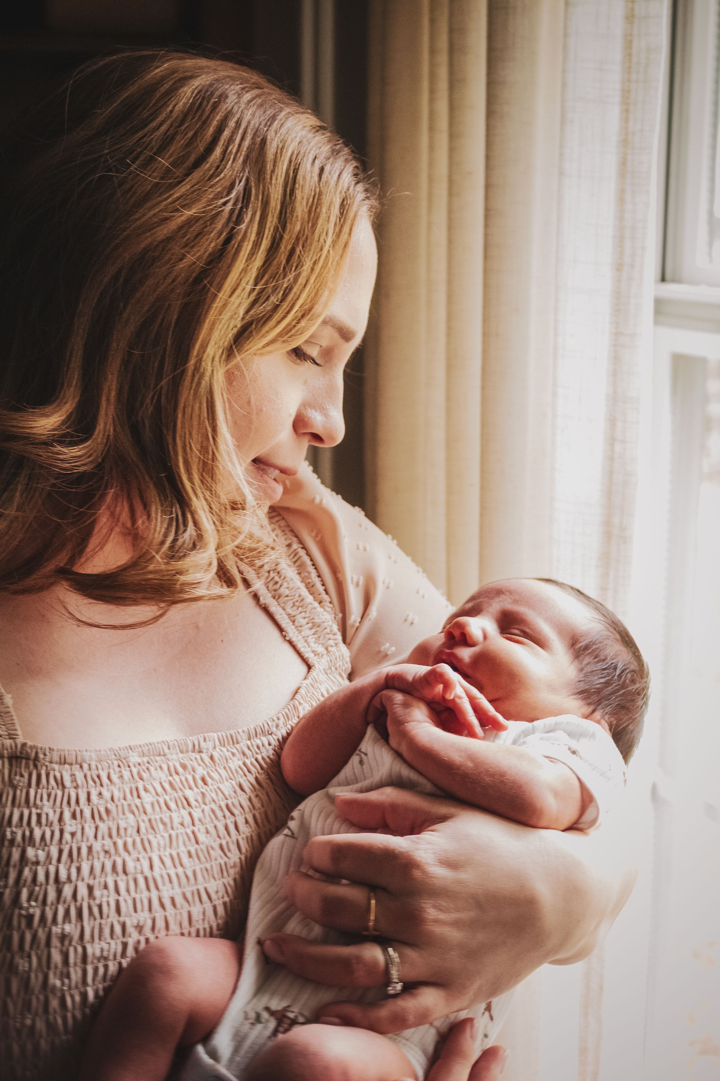 A smiling mother tenderly holds her sleeping newborn baby near a sunlit window.