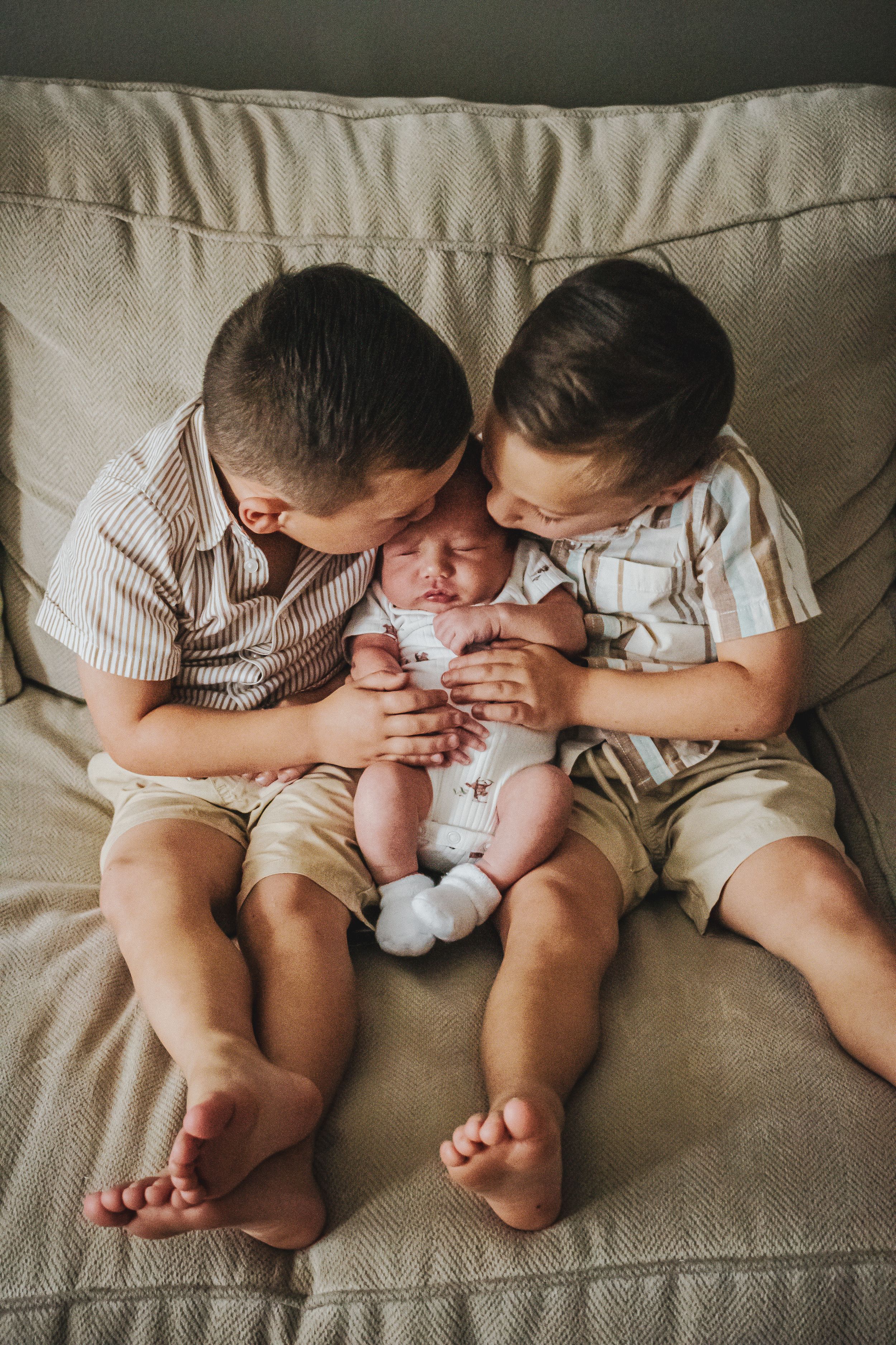 Two young boys sitting on a couch lovingly holding and kissing a newborn baby sibling.