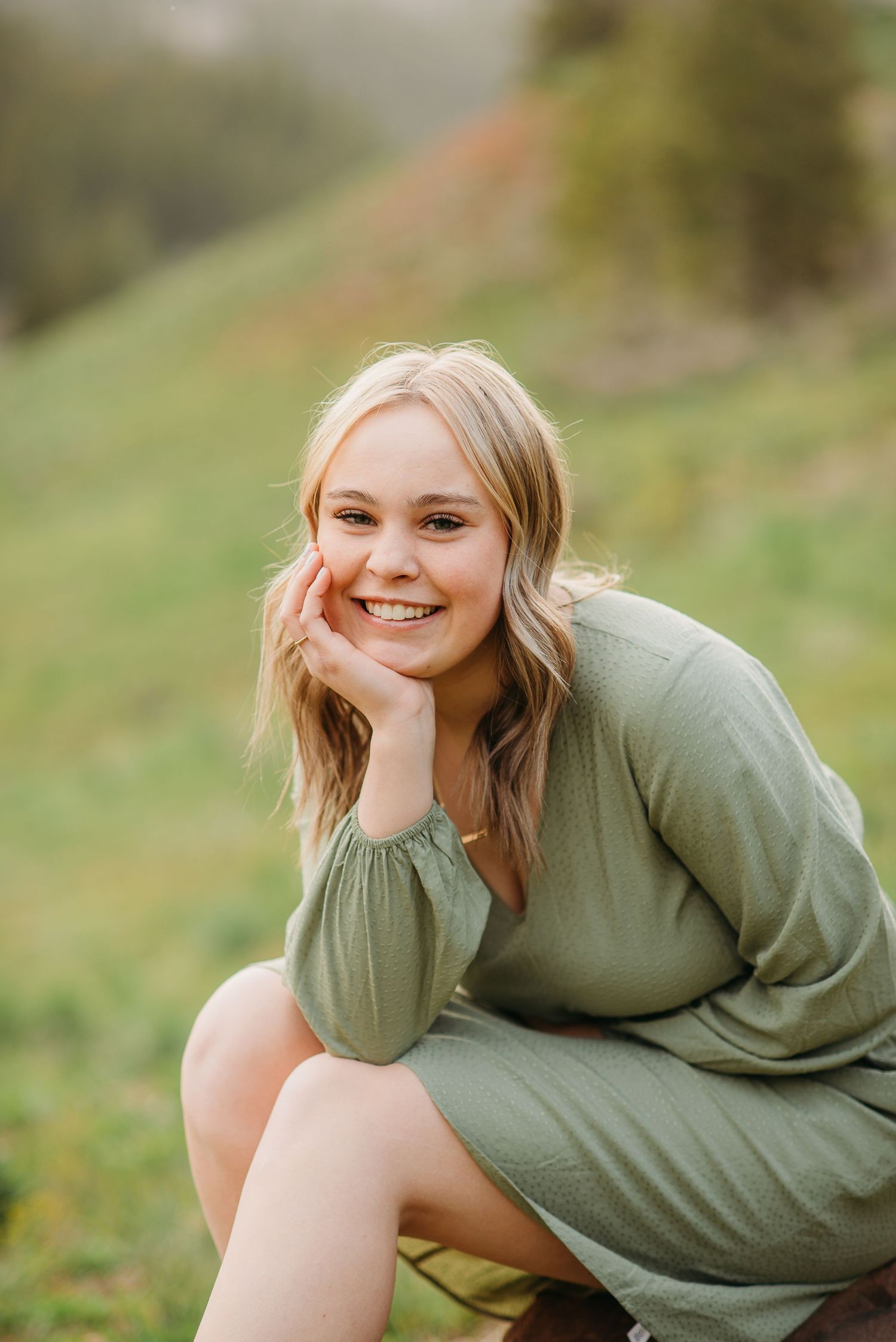 Young woman in sage green dress sits casually outdoors on grassy hillside.