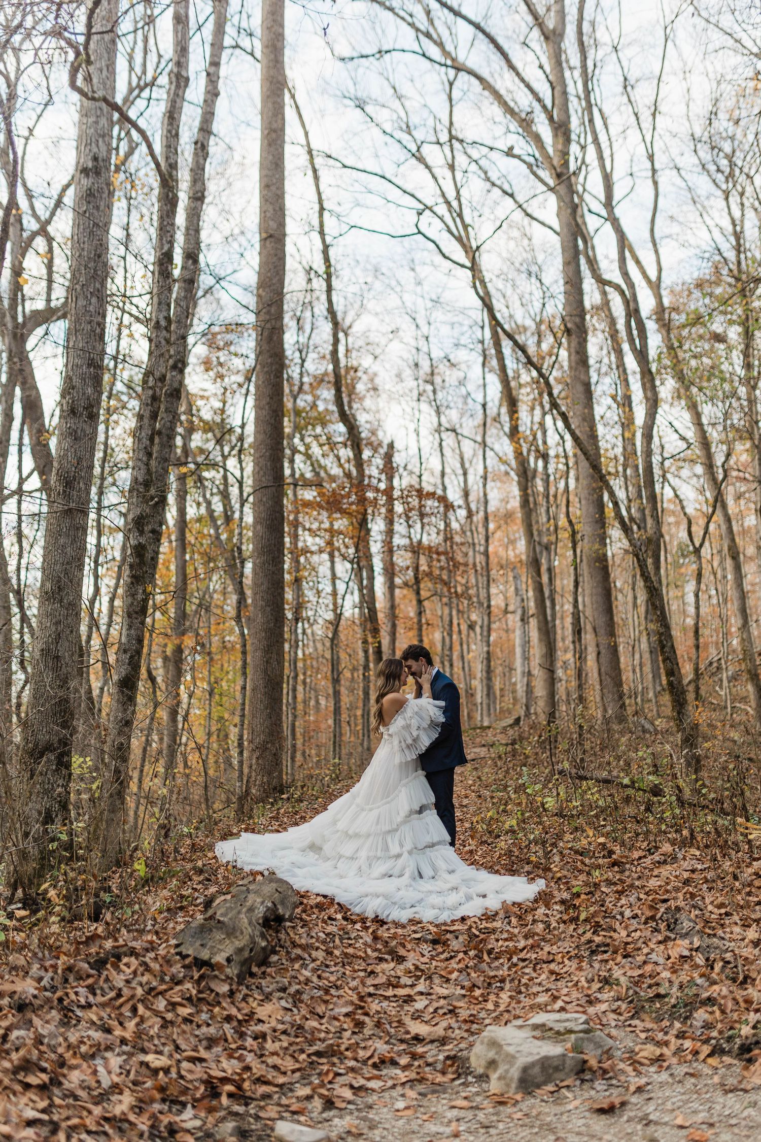 A couple in formal wedding attire embrace in a forest path covered with fallen autumn leaves.