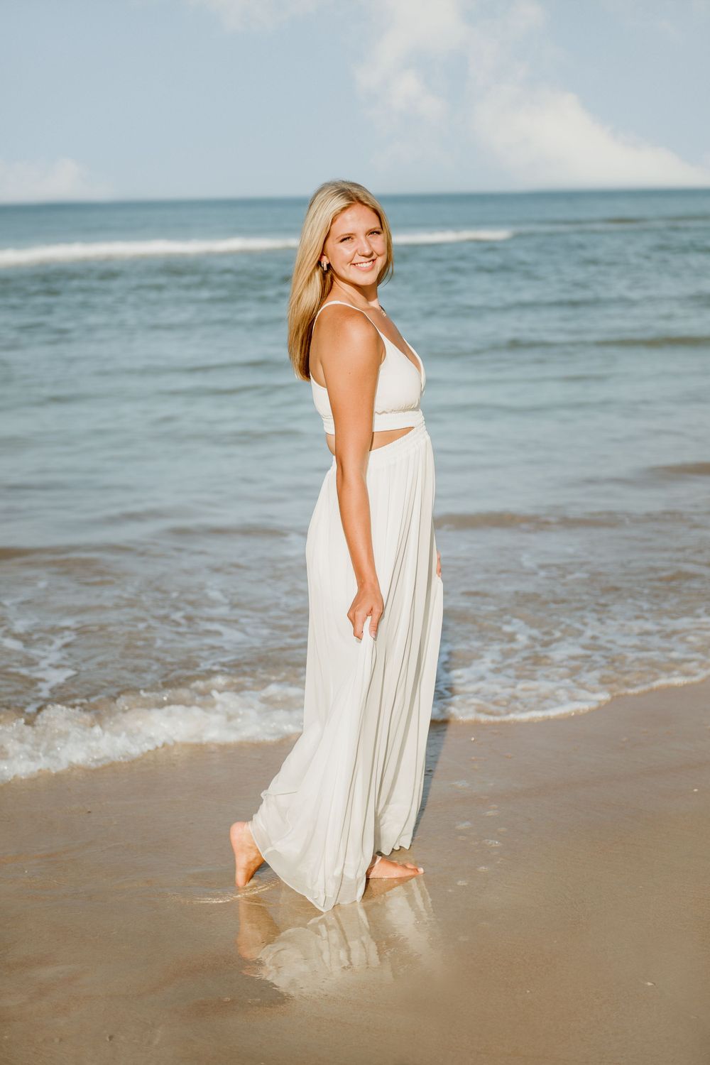 Woman in flowing white dress stands at ocean's edge during sunset.