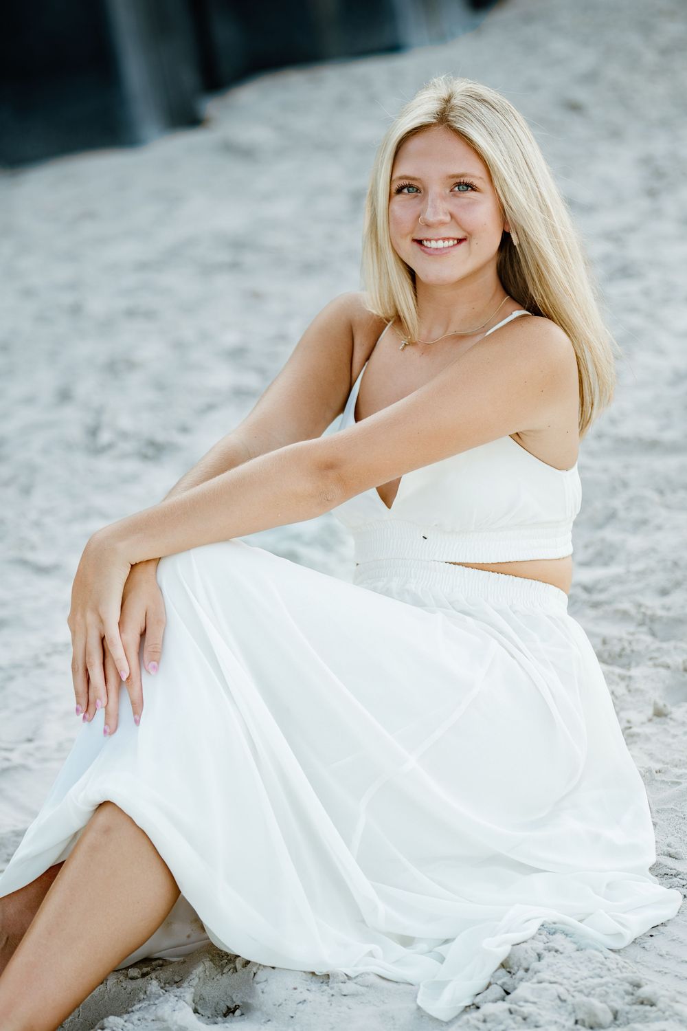 Woman in white outfit sits gracefully on sandy beach.
