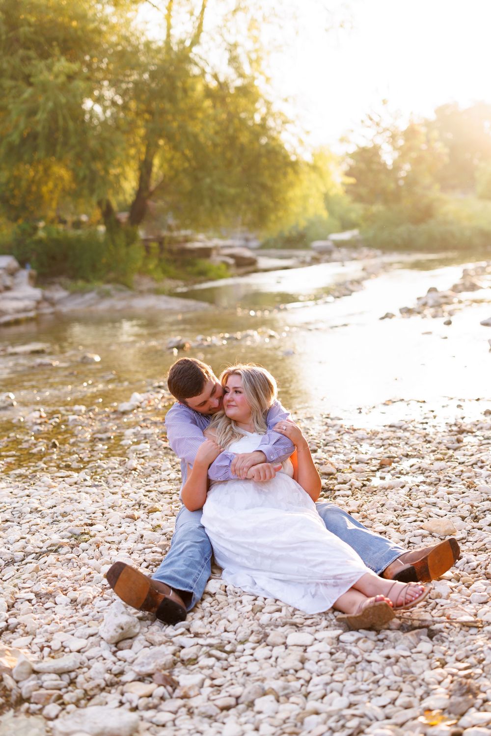 A romantic photoshoot of a couple embracing on a rocky riverbank at sunset.