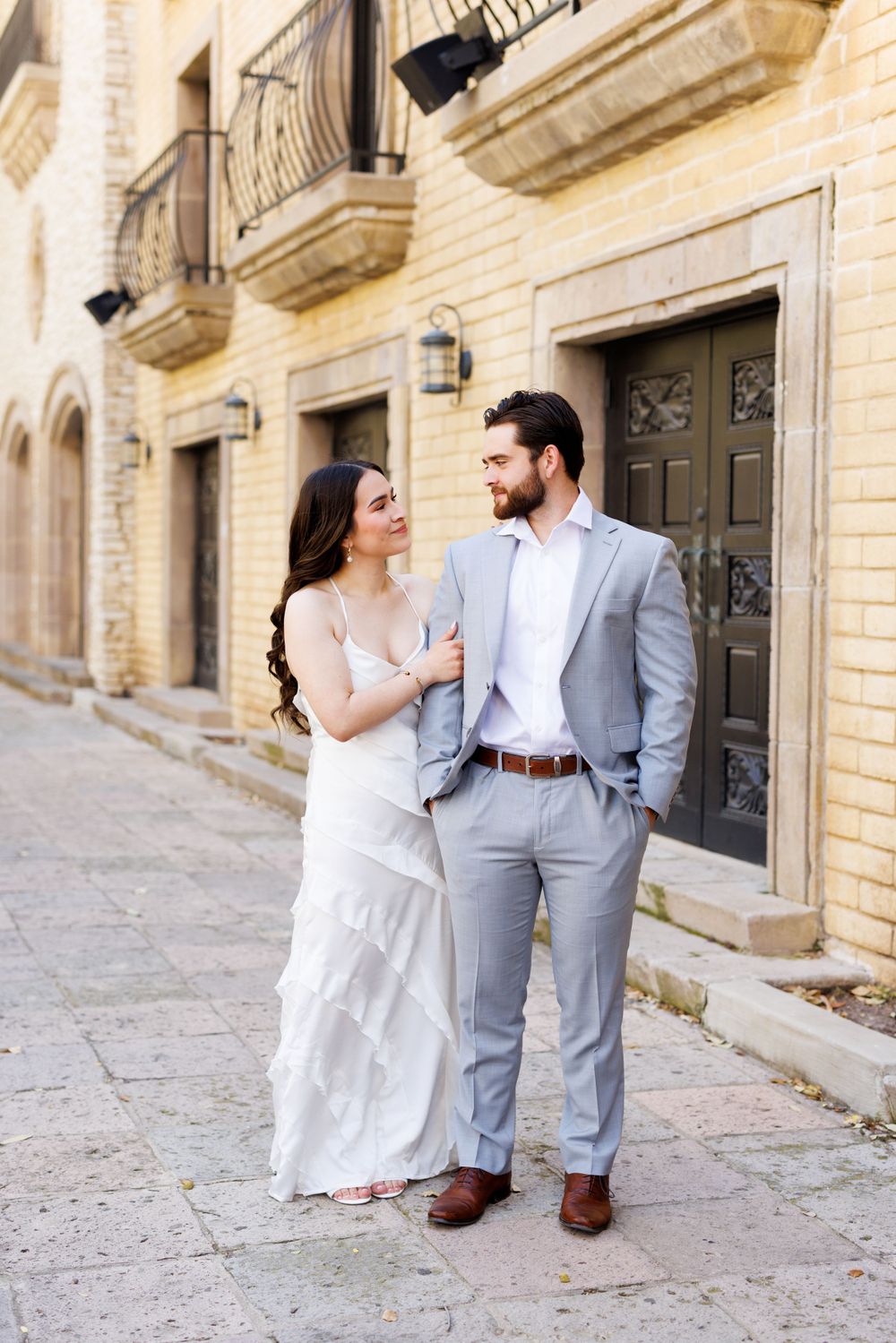 Romantic couple in white dress and gray suit pose against yellow brick wall in elegant engagement photos.