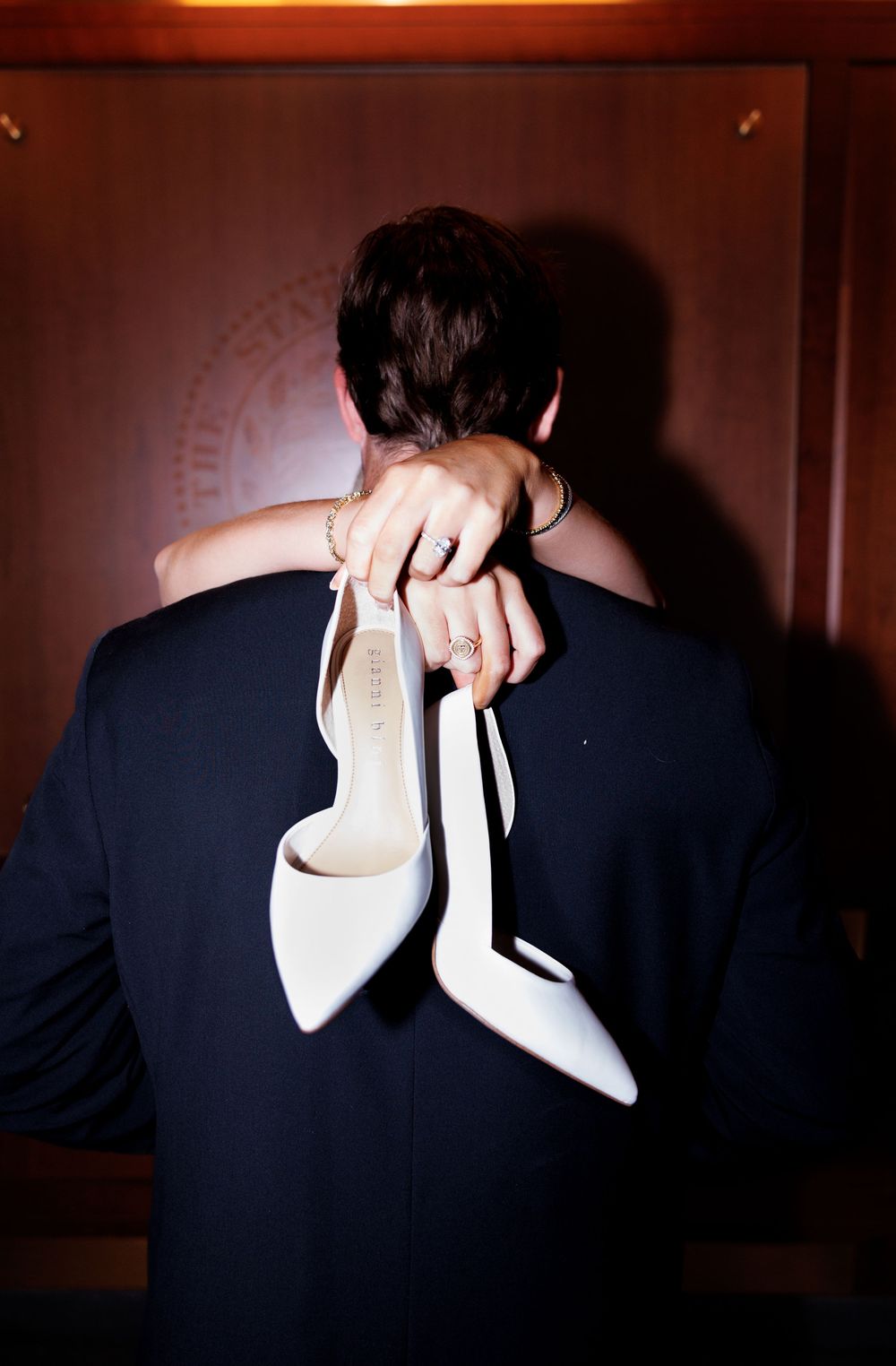A person in a suit holds white high heels while embracing someone in a white dress.