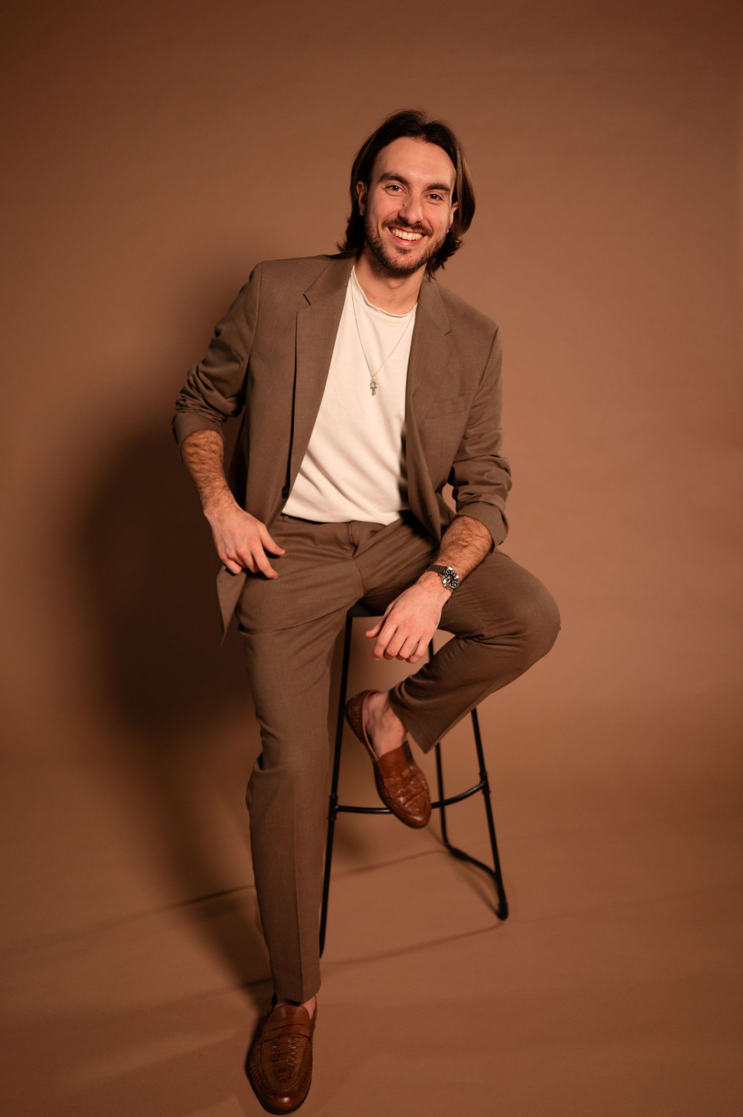 portrait of bon amour weddings owner Jordan sitting on a chair posing wearing a brown suit and beige t shirt
