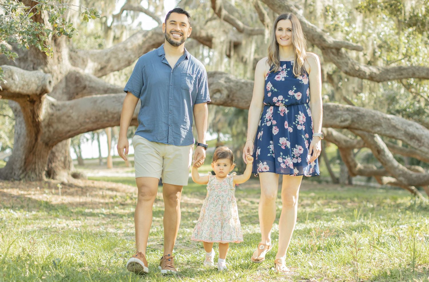 Family holding hands while standing together under Spanish moss draped trees in an outdoor garden setting.