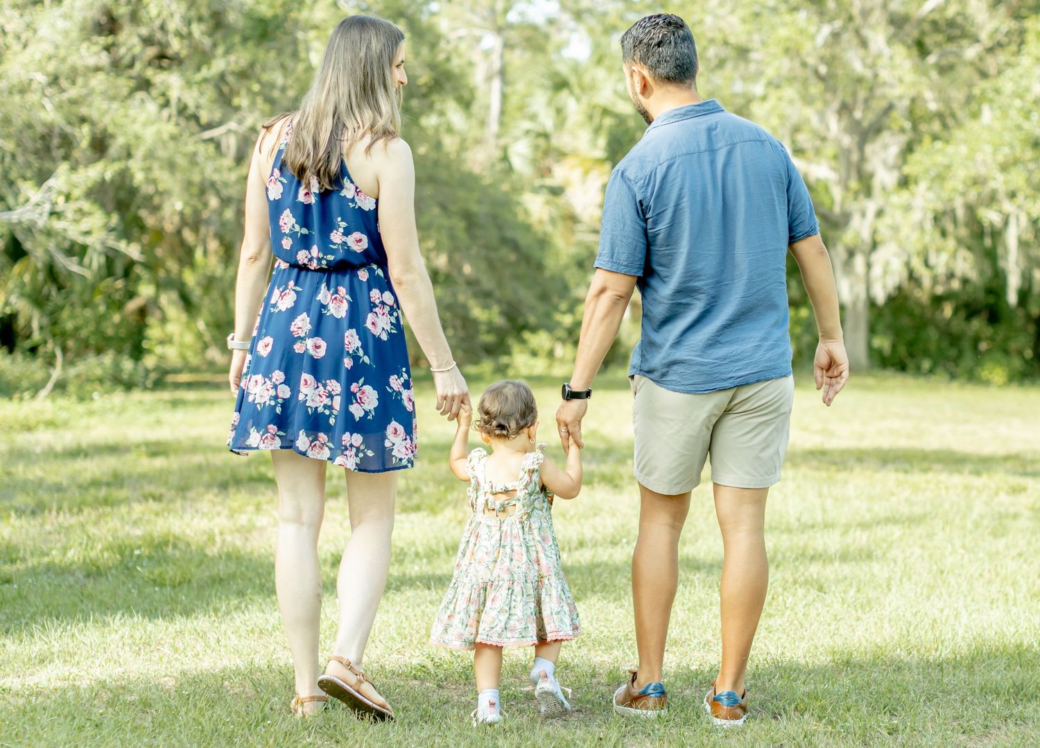 A family holds hands while walking together through a sunny park, wearing blue and floral summer outfits.