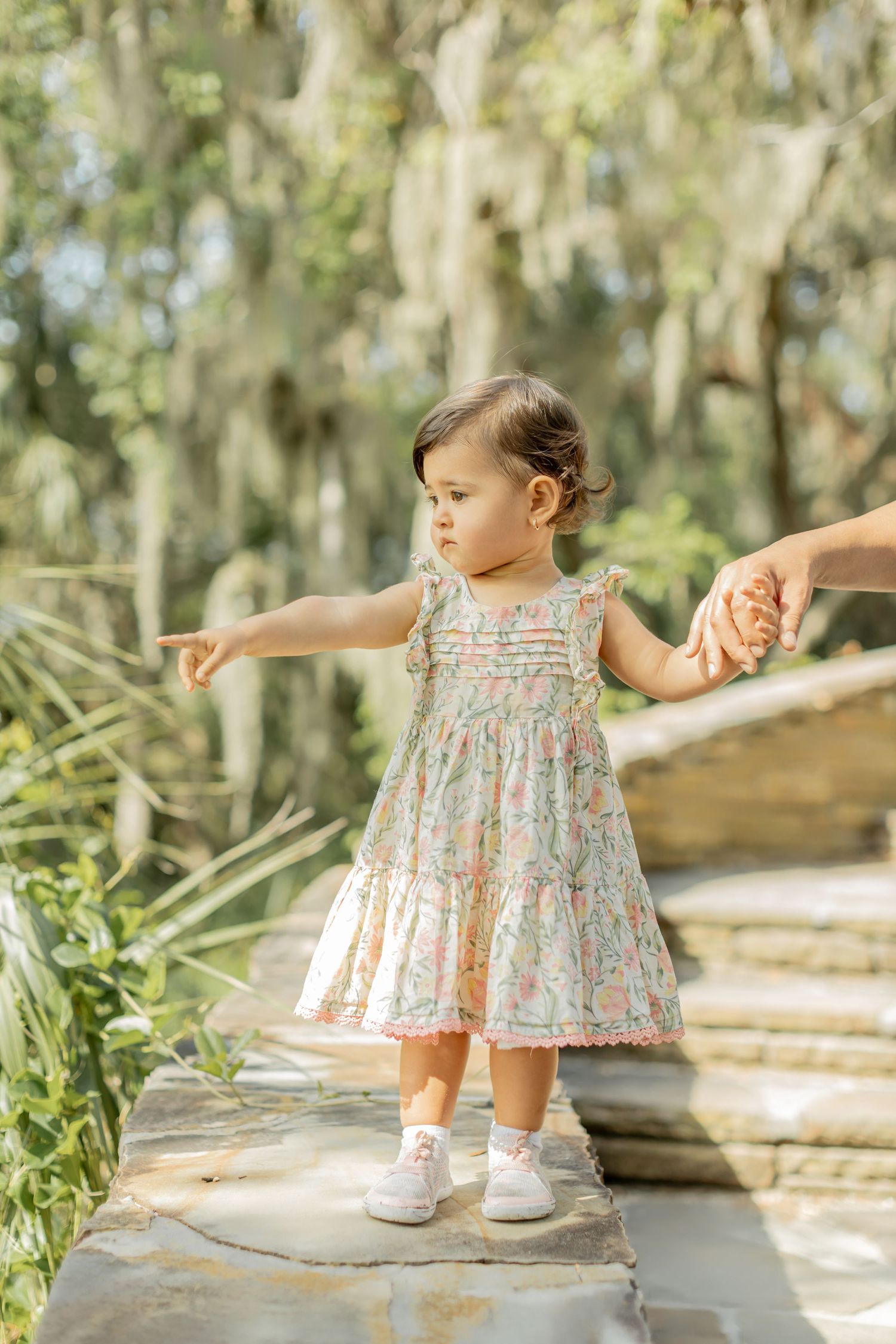 A small child in a floral dress stands on a stone path in a serene garden setting with Spanish moss in background.