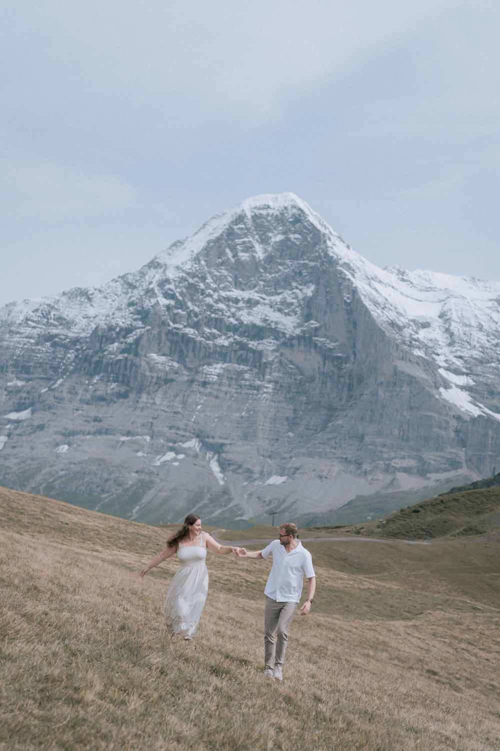 A couple in white outfits runs playfully along a grassy hillside with snow-capped Swiss Alps mountains in the background.