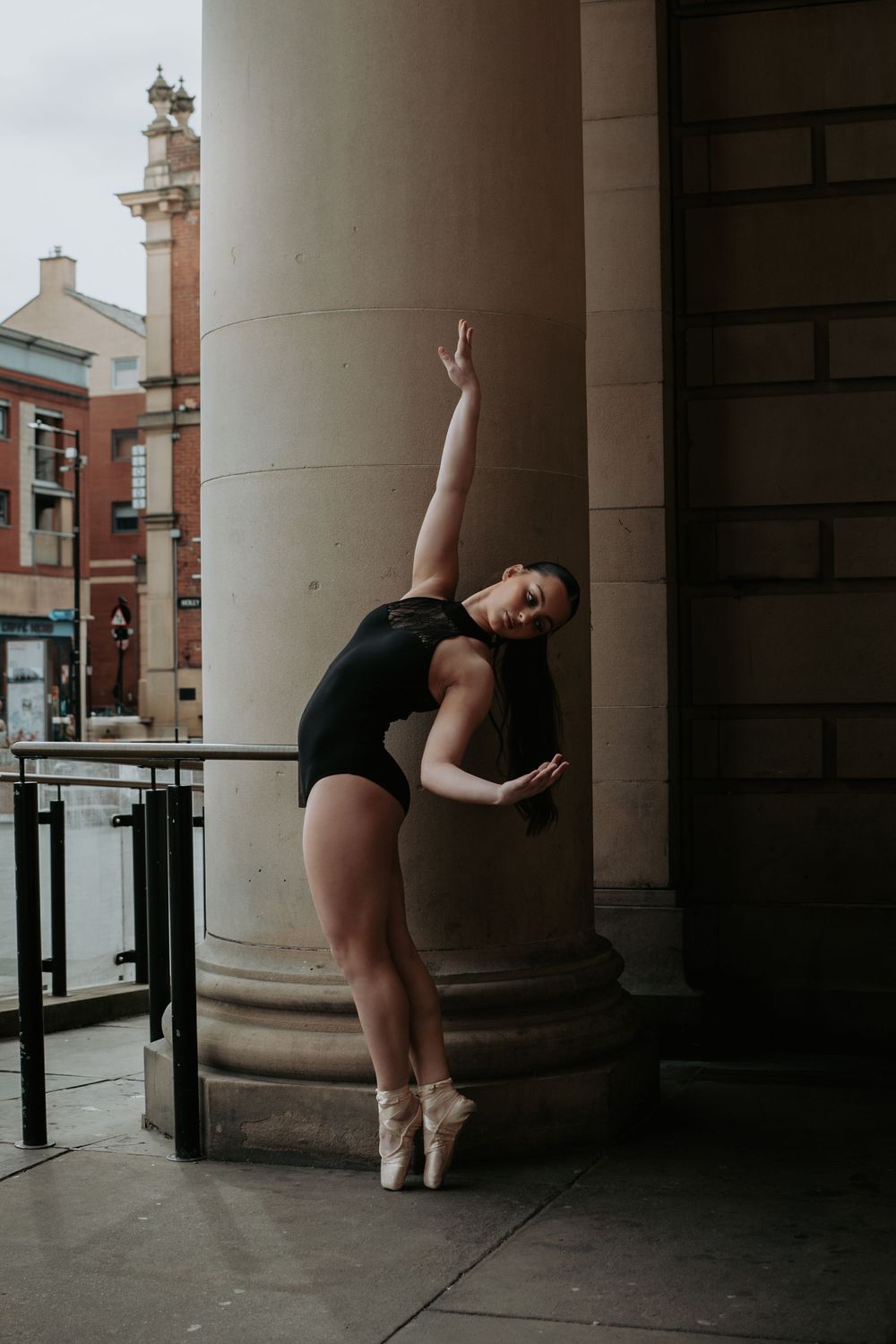 A ballet dancer in black leotard performs graceful poses against architectural columns in an urban setting.