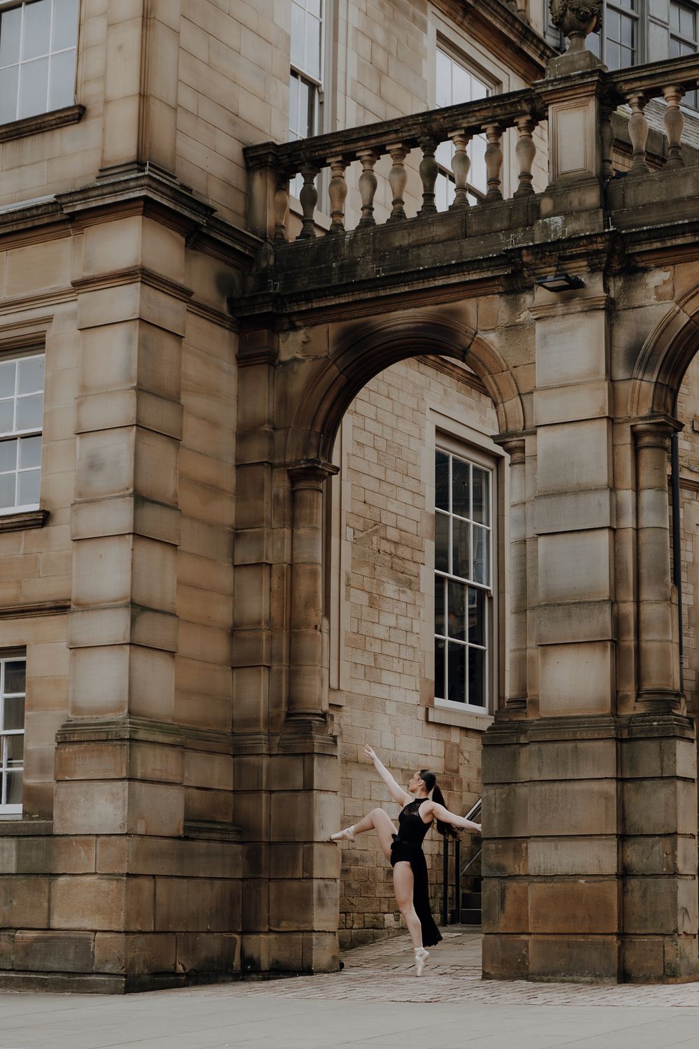 A dancer performs a graceful movement against a historic stone building facade with arched windows and columns.