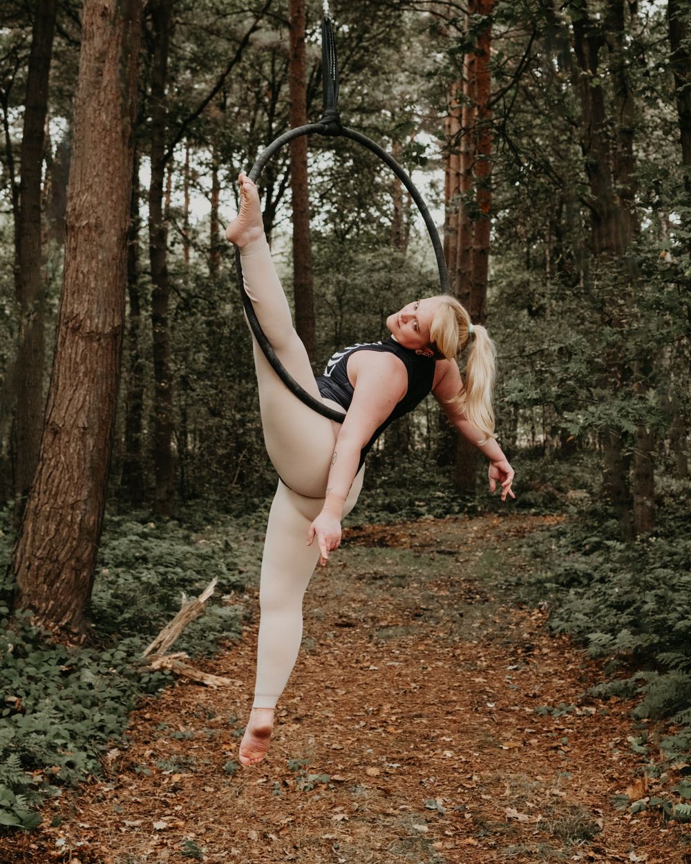 Dancer performs elegant high leg extension pose on forest trail during artistic outdoor photoshoot.
