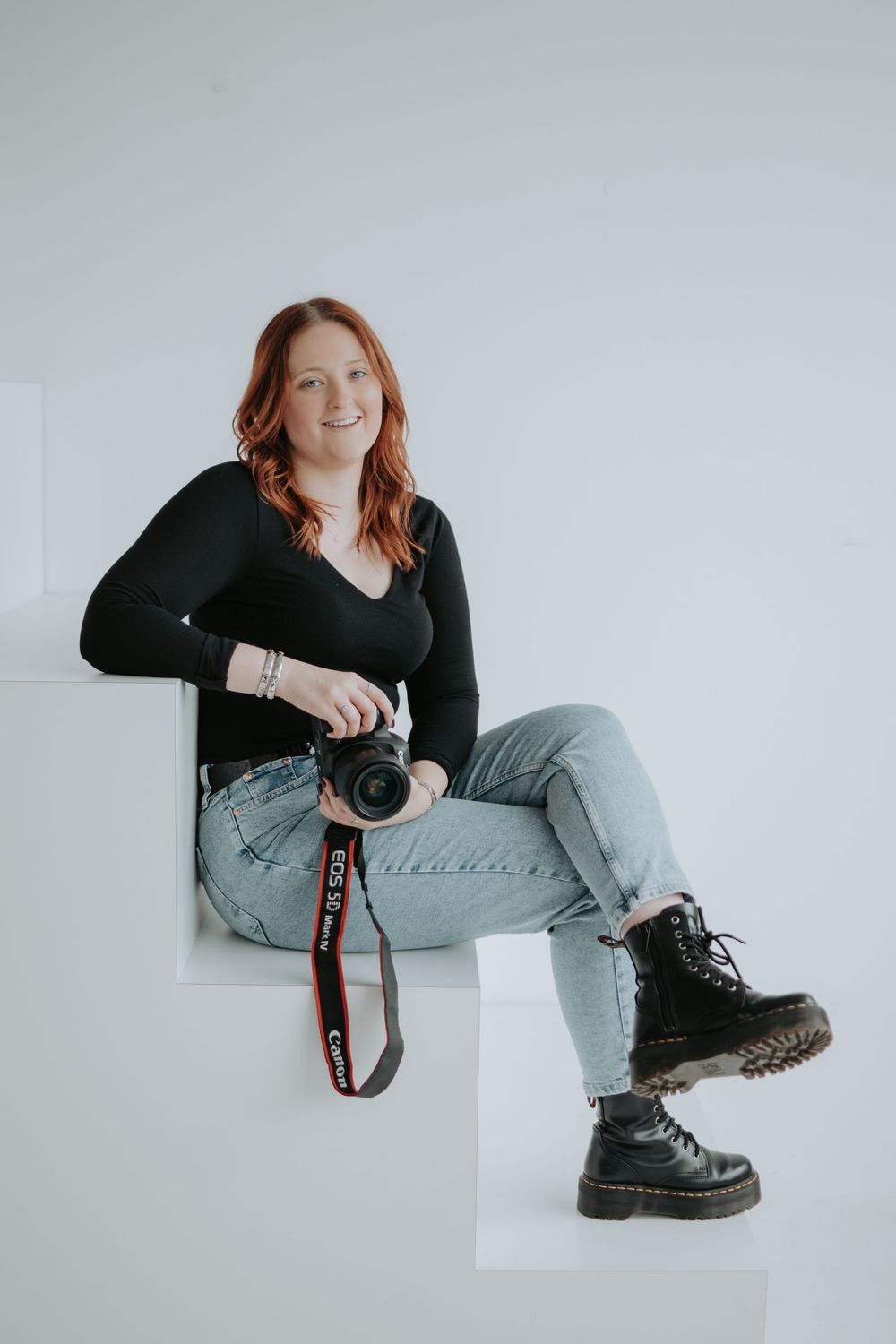Photographer in black sweater and jeans sits casually on white steps while holding professional camera.