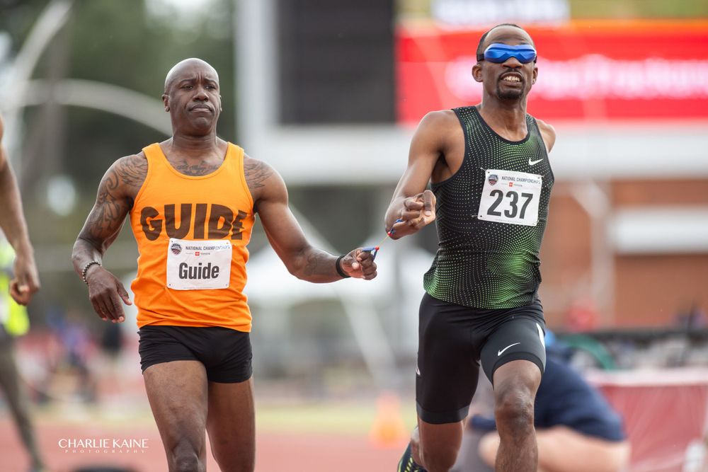 Two athletes competing together with one wearing orange guide vest during track event.