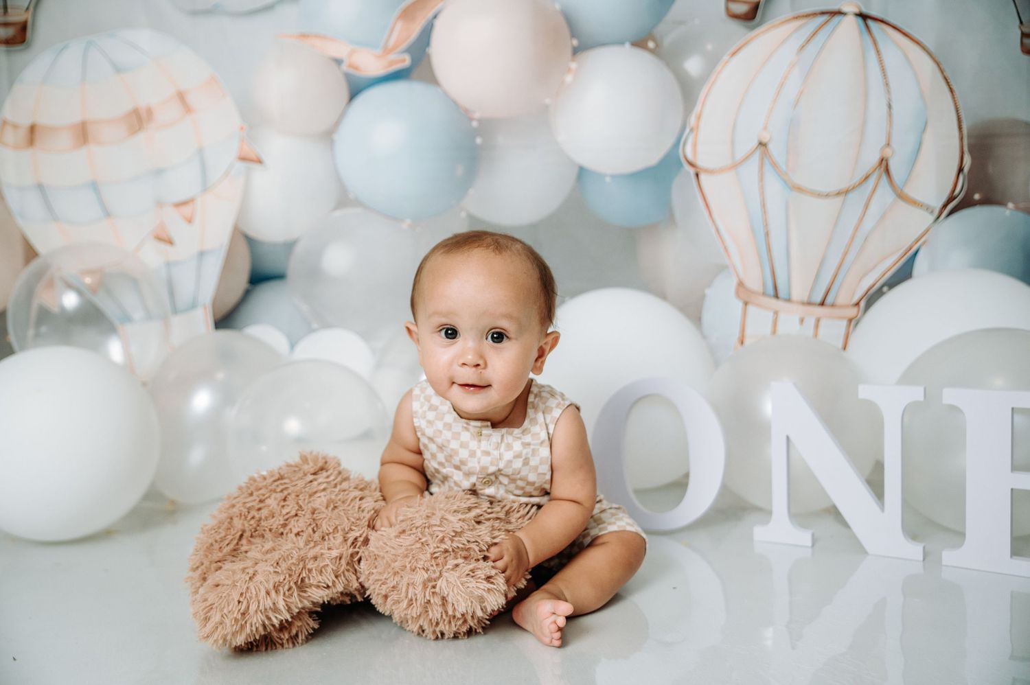 Baby sitting with teddy bear surrounded by white and blue balloons with hot air balloon decorations.
